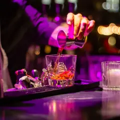 Barman nalewa bursztynowy cordial drink do szklanki z lodem. W tle kolorowe światła.